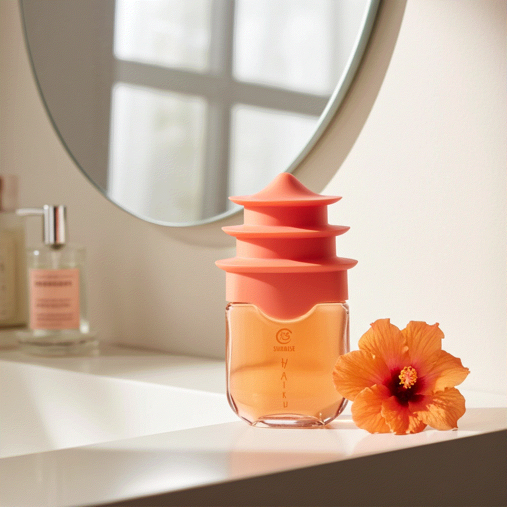 Haiku Sunrise Eau De Perfume 1.7 fl oz bottle with orange floral cap and hibiscus flower on white countertop under mirror
