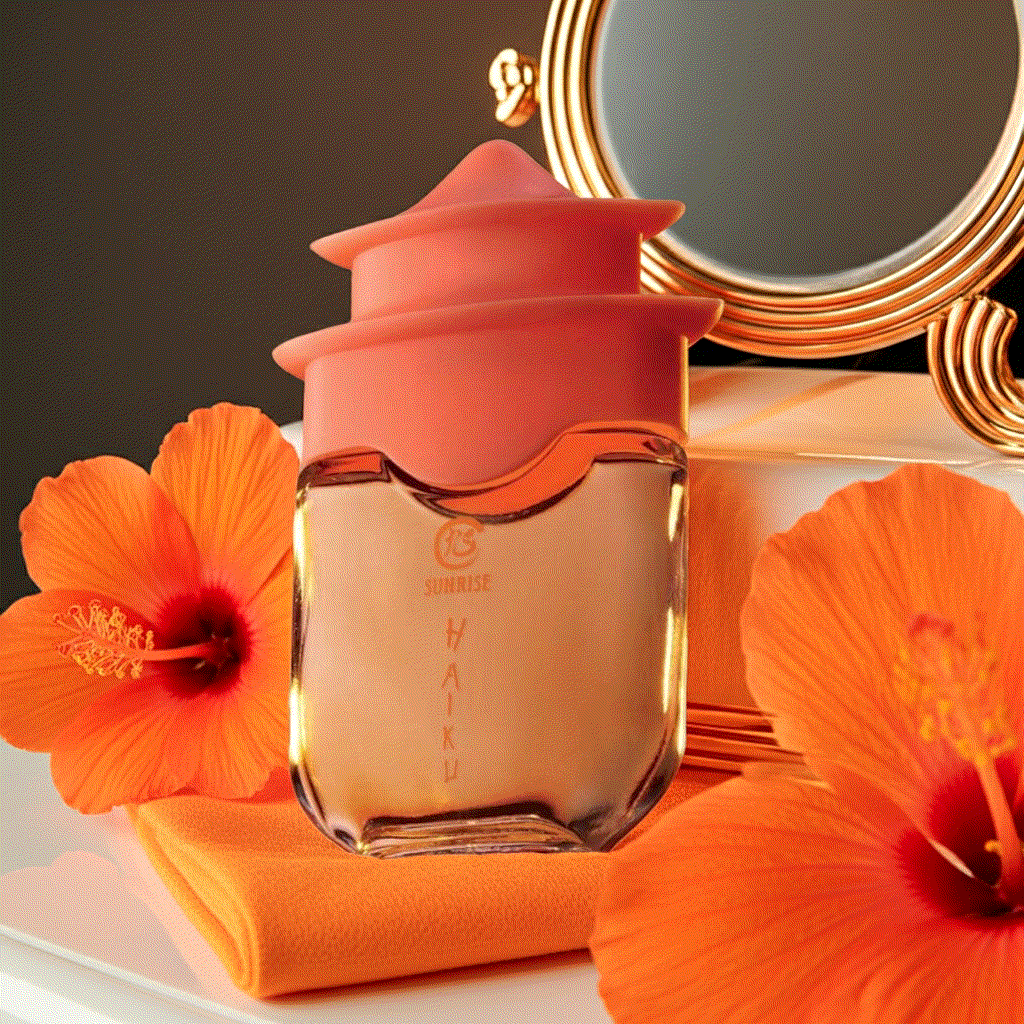 Haiku Sunrise Eau De Perfume bottle with orange floral cap and hibiscus flowers on orange cloth near gold mirror