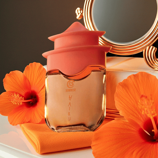 Haiku Sunrise Eau De Perfume bottle with orange floral cap and hibiscus flowers on orange cloth near gold mirror